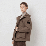 A woman with her hair pulled back, wearing a brown heated assault coat with a hood and multiple pockets, including a black patch on the left sleeve. She is looking towards the viewer.