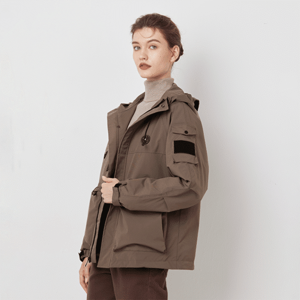 A woman with her hair pulled back, wearing a brown heated assault coat with a hood and multiple pockets, including a black patch on the left sleeve. She is looking towards the viewer.