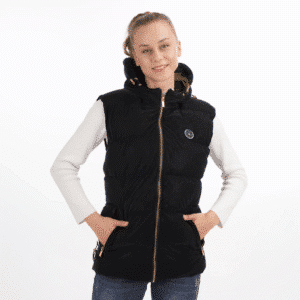 A young woman with her hair tied back, smiling and standing with her hands in the pockets of a black hooded heated vest, worn over a white long-sleeved shirt and blue jeans.