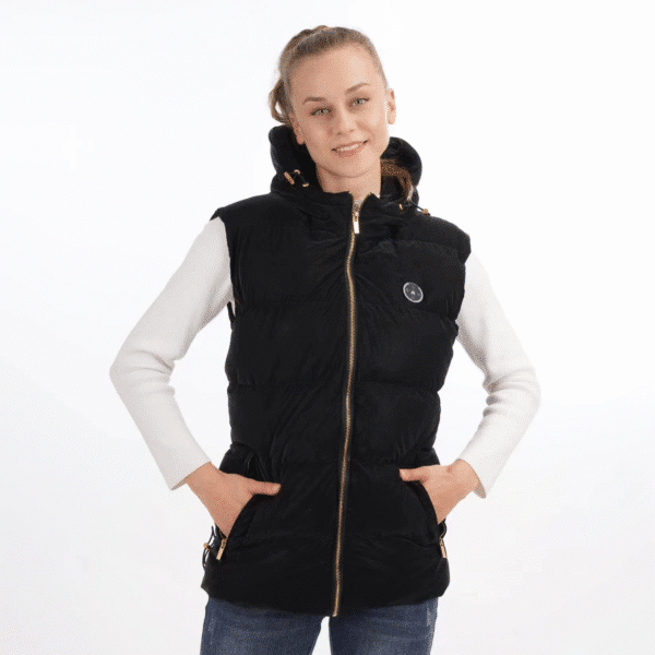 A young woman with her hair tied back, smiling and standing with her hands in the pockets of a black hooded heated vest, worn over a white long-sleeved shirt and blue jeans.