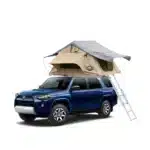 A dark blue Toyota 4Runner SUV with a light brown and grey roof top tent mounted on its roof rack, with a silver ladder extending to the ground.